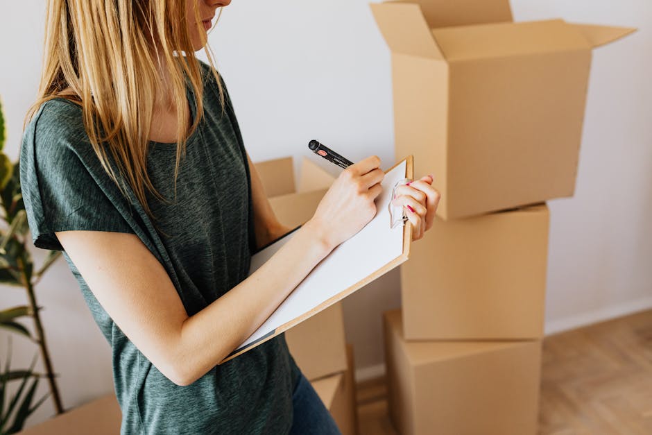 A woman with long blonde hair, wearing a dark green short-sleeved shirt, is holding a clipboard and writing with a black marker. She is indoors, surrounded by several large cardboard moving boxes, som