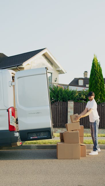 A woman with long blonde hair, wearing a dark green short-sleeved shirt, is holding a clipboard and writing with a black marker. She is indoors, surrounded by several large cardboard moving boxes, som