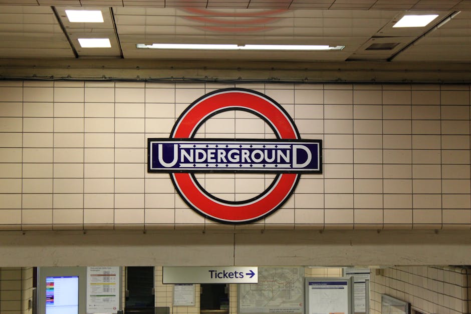 A large circular London Underground sign with a red outer ring and a blue inner rectangle displaying the word 'UNDERGROUND' in white, situated on a tiled wall inside a station. Below the sign, there i